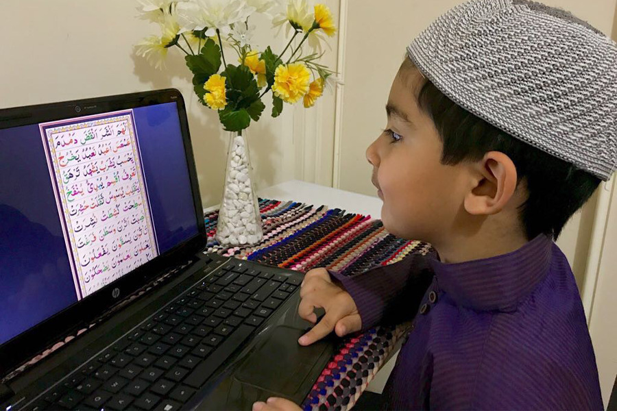 Young boy in purple shirt pointing at a laptop screen with Quranic text